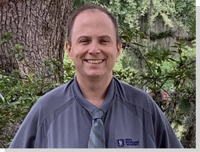 A man in a blue shirt and tie standing next to a tree.