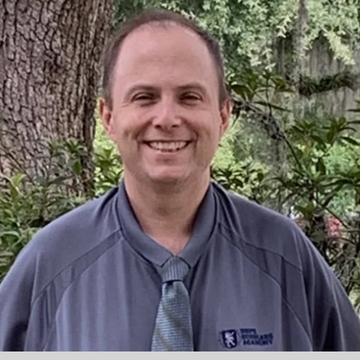 A man in a blue shirt and tie standing next to a tree.